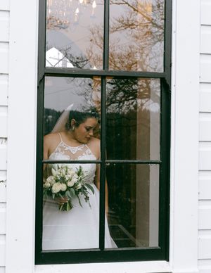 A bride in a white gown stands by a window holding a bouquet while looking down.
