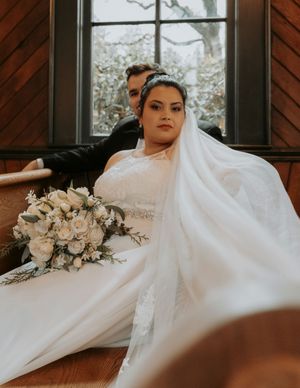 A bride in a flowing white wedding dress poses elegantly on a wooden floor with a romantic floral bouquet nearby.