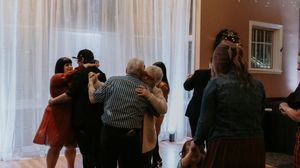 Couples dancing together at an indoor social gathering with white curtains in the background.