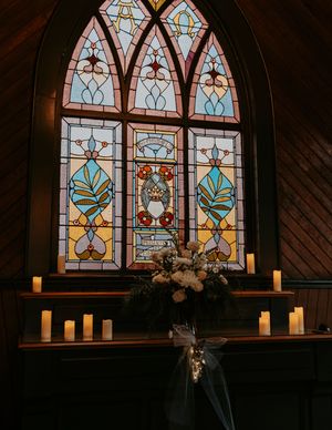 A series of stained glass church windows with candles lit beneath them creating a warm, atmospheric glow.