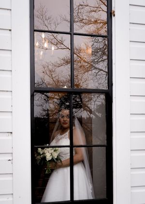 Through a window pane, a bride in a white wedding dress is seen holding a bouquet against white shutters.