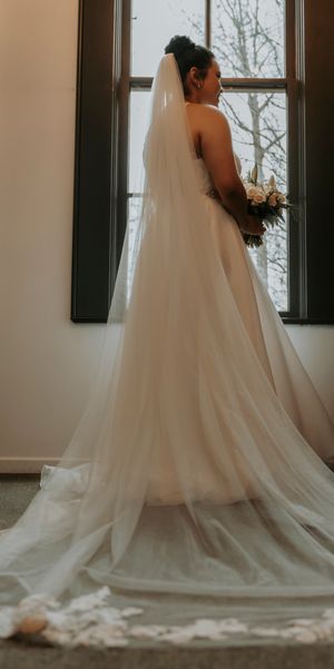 A bride in a flowing white wedding gown with a cathedral-length veil stands elegantly against a light interior backdrop.