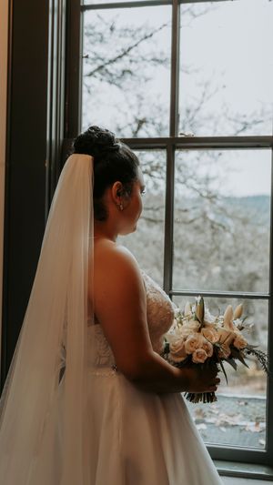 A bride in a white gown gazes out a window while holding a bouquet of light-colored flowers.