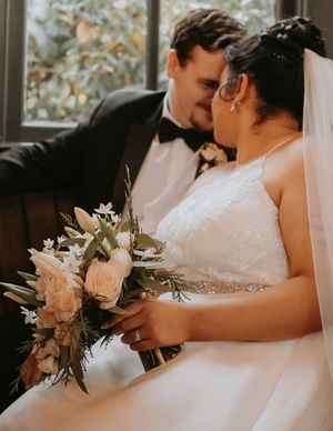 Romantic wedding moment with couple in formal attire holding a blush and white floral bouquet by window.