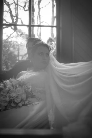 A dreamy black and white bridal portrait by a window with flowing veil and bouquet creating an ethereal atmosphere.