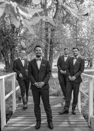 Groomsmen in black suits pose playfully on a white wooden bridge in a natural outdoor setting.