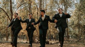 A group of groomsmen in dark suits jump in unison during a fall wedding photo session in a wooded setting.
