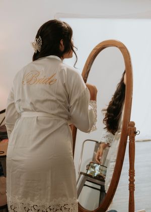 A white robe with 'Bride' embroidery hangs on a wooden hanger against a light background.