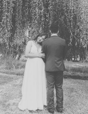 A couple in wedding attire stands together under a weeping willow tree in a romantic black and white photograph.