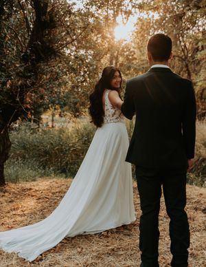 A romantic sunset wedding photo showing a couple in formal attire on a wooded path with golden light filtering through trees.