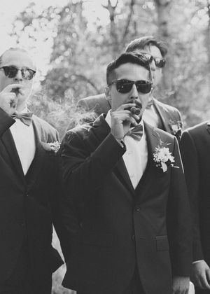 Black and white photo of groomsmen in tuxedos and sunglasses smoking cigars during wedding celebrations.