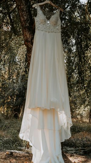 An elegant white wedding dress with lace details hangs from a tree branch in natural sunlight.