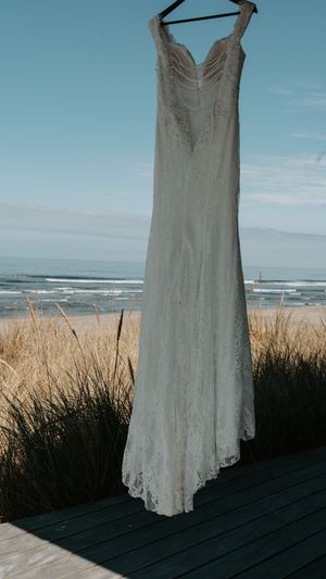 An elegant white lace wedding dress hangs against a coastal backdrop with beach grass and ocean waves.