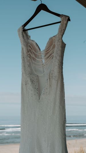 A white lace wedding dress hangs on a hanger against a beach backdrop with ocean waves and blue sky.