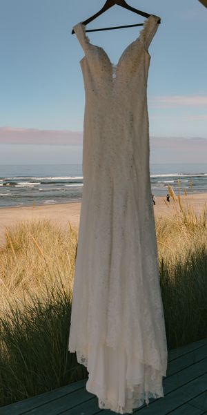 A white lace wedding dress hangs against a coastal backdrop with beach grass and ocean waves.