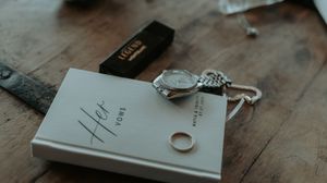 A silver ring and ornate key rest beside a gray book on a weathered wooden surface.