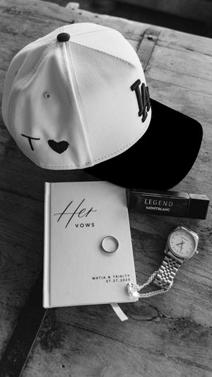 Black and white image of wedding accessories including a white baseball cap and vows book on a wooden surface.