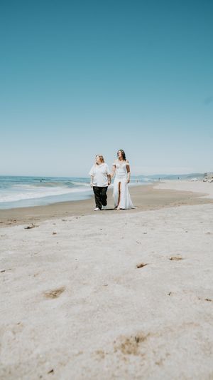 People in white dresses walking with a black dog along a serene beach with blue sky and ocean in the background.