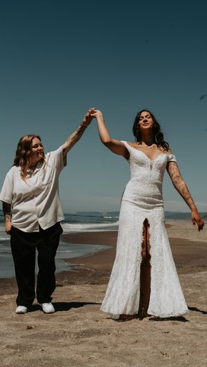 Two people in white outfits posing joyfully on a sandy beach against a blue sky backdrop.