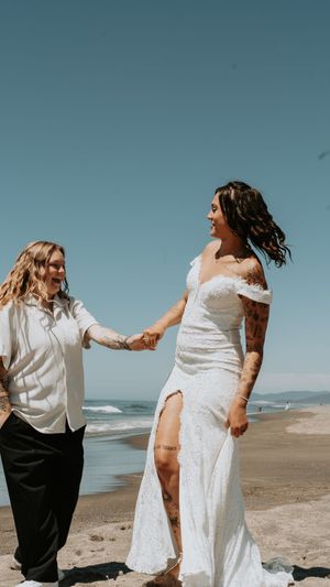 Two people in white outfits dance joyfully together on a sunny beach with ocean waves in the background.