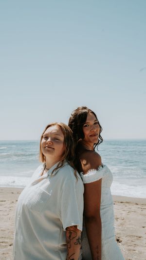 Two friends in white outfits share joyful moments together on a sunny beach with ocean waves in the background.