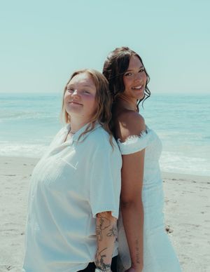 Two friends in white outfits pose back-to-back on a sunny beach with ocean waves in the background.