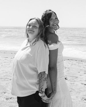 Two people stand back-to-back smiling on a sunny beach in black and white.
