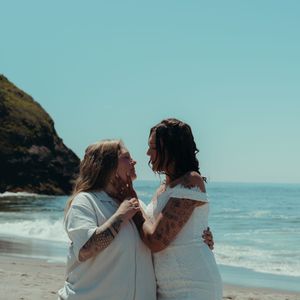 Two people in white outfits share intimate moments at a scenic beach with cliffs and ocean waves in the background.