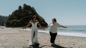 A couple in white clothing walks together on a sandy beach with ocean waves in the background.