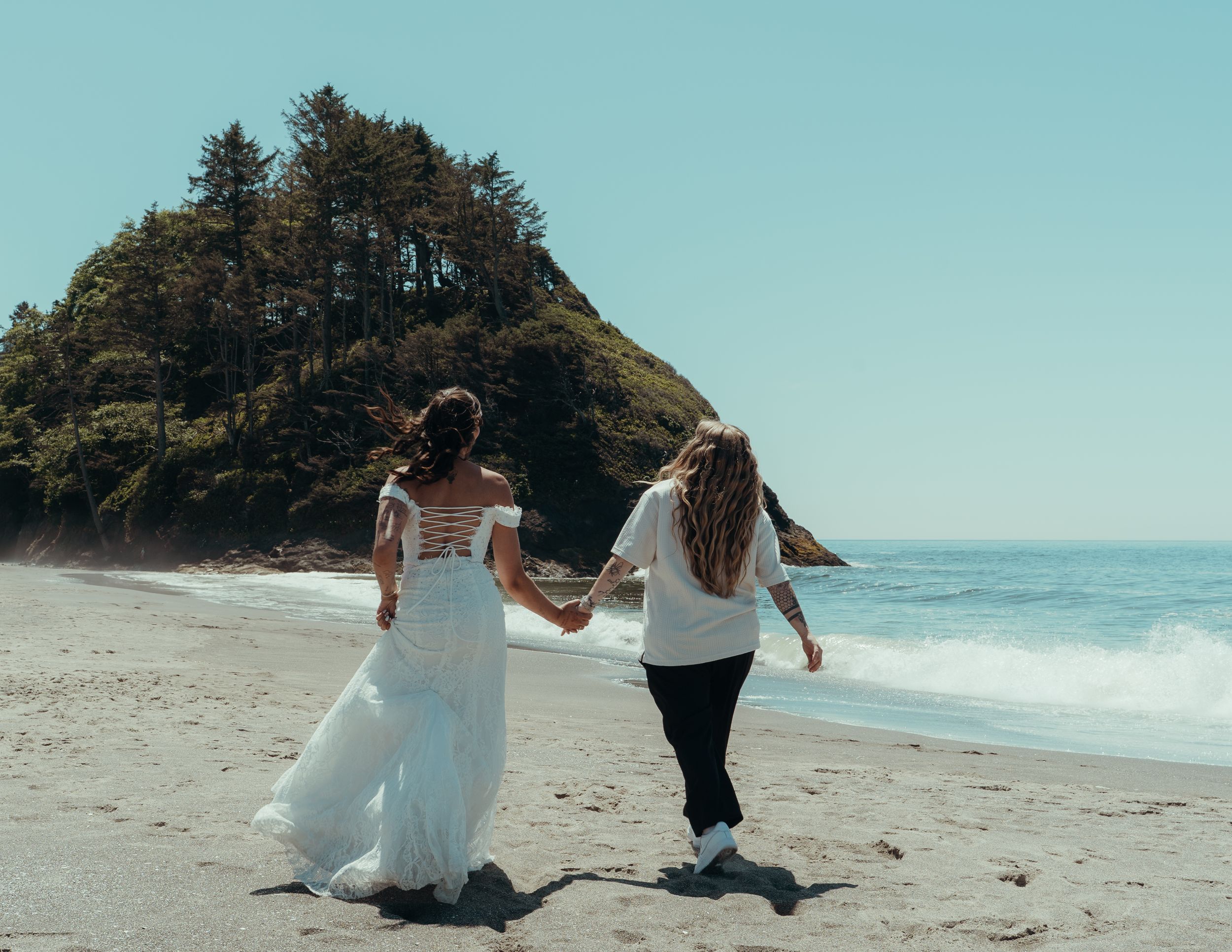 Two people in wedding attire walk hand in hand along a scenic beach with waves crashing and trees in the background.