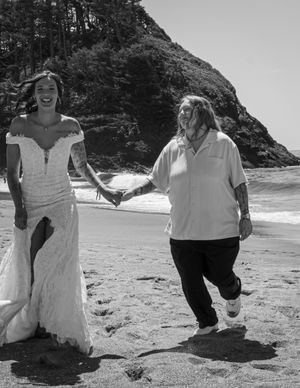 A group of people in white clothing run and play joyfully on a scenic beach with rocky cliffs in black and white.