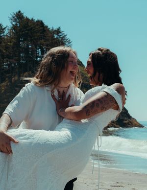 A joyful couple in wedding attire share a laugh while posing on a scenic beach with evergreen trees and ocean views.