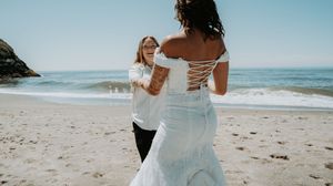 Panoramic beach wedding scene with guests gathered along the shoreline under blue skies.