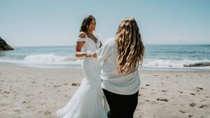 A panoramic beach scene shows two people in contrasting white and black outfits embracing by the ocean.