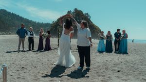 Wide angle view of a beach ceremony with attendees in formal wear gathered by the ocean.