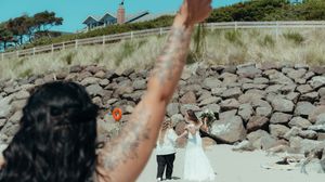A couple celebrates their wedding day near a rocky seawall with a lighthouse visible in the distance.