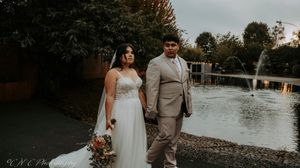 A couple celebrates their wedding day in a rustic outdoor setting with a waterfront backdrop at sunset.