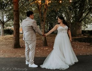 A couple in wedding attire walks together on a tree-lined path with twinkling lights overhead during sunset.