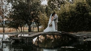 A wedding couple stands by a serene lake surrounded by autumn trees creating a romantic reflection on the water.