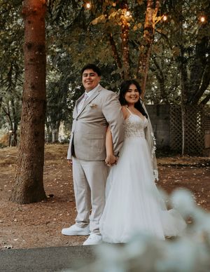 Newlyweds in white attire walking hand in hand through a scenic wooded area with warm lighting.