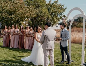 Outdoor wedding ceremony under white arch with bridesmaids in dusty rose dresses and groomsmen in light suits.