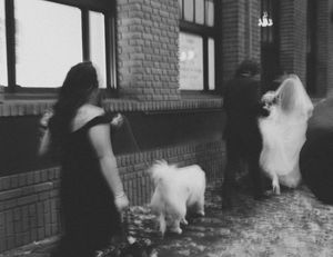 A woman in a dark dress stands with two white dogs outside a brick building in a vintage black and white photo.