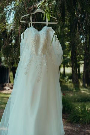 A delicate white wedding dress with lace details hangs from a hanger against a green outdoor background.