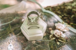 A delicate engagement ring sits in a hexagonal ring box surrounded by scattered flower petals and greenery.