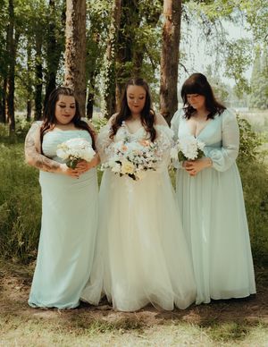 Bridesmaids in soft mint green dresses and white bouquets pose together in a wooded outdoor setting.