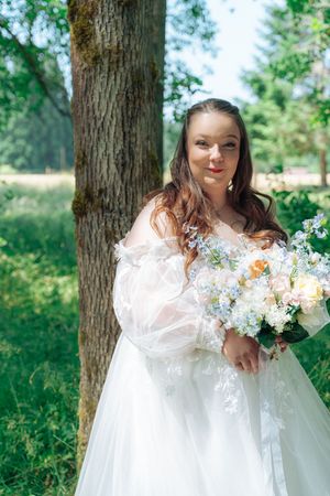 A bride in a white lace gown holds a delicate bouquet of white and peach flowers in a sunlit forest setting.