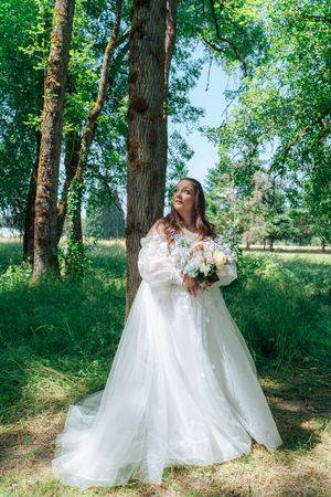 A dreamy bridal photoshoot in a sunlit forest shows flowing white wedding gowns against lush green trees.