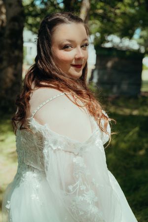 A bride in a delicate white lace wedding dress poses outdoors in a sunlit garden setting.