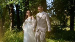 A couple in white wedding attire walks together through a sunlit wooded area on a summer day.