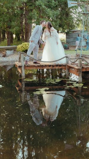 A romantic wedding scene reflects in still pond water with a rustic wooden bridge and trees in the background.